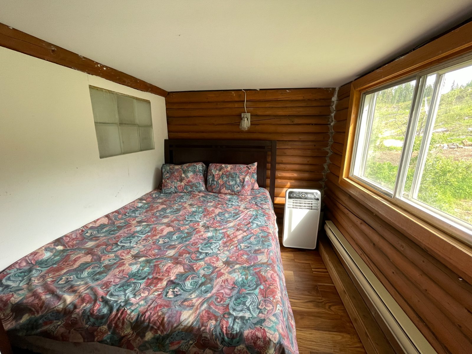 The second bedroom with a bed against the warm log wall, a portable air conditioner, and a wide window
